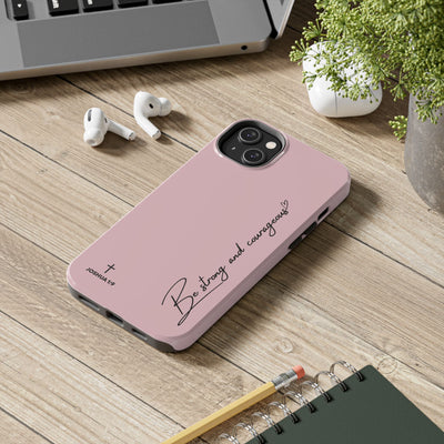 Pink inspirational tough phone case with "Be strong and courageous" scripture design on wooden desk with earbuds and notebook