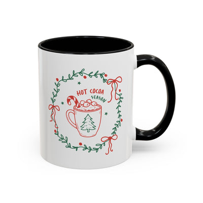 white ceramic mug with black handle and interior featuring hot cocoa season holiday design and greenery wreath