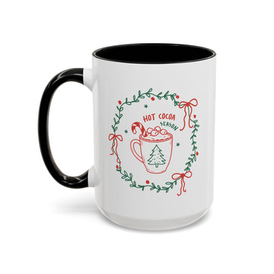 White ceramic mug with black handle and interior, festive hot cocoa season design with greenery wreath, red and green accents
