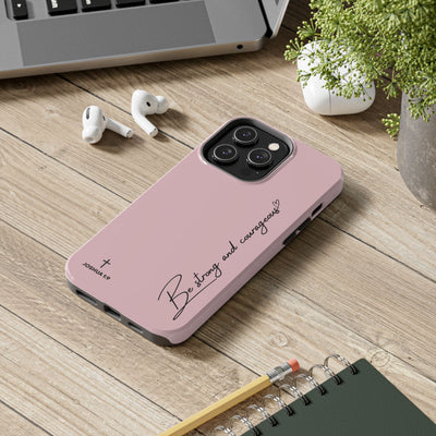 Inspirational tough phone case in pink with "Be strong and courageous" text and Joshua 1:9 verse on a wooden desk with earbuds and notebook