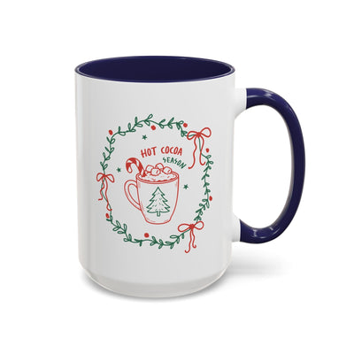 White ceramic mug with navy interior and handle, featuring festive "Hot Cocoa Season" design with greenery wreath and holiday motifs