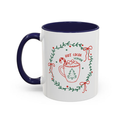 white ceramic mug with navy handle and interior featuring hot cocoa season design with greenery wreath and red bows